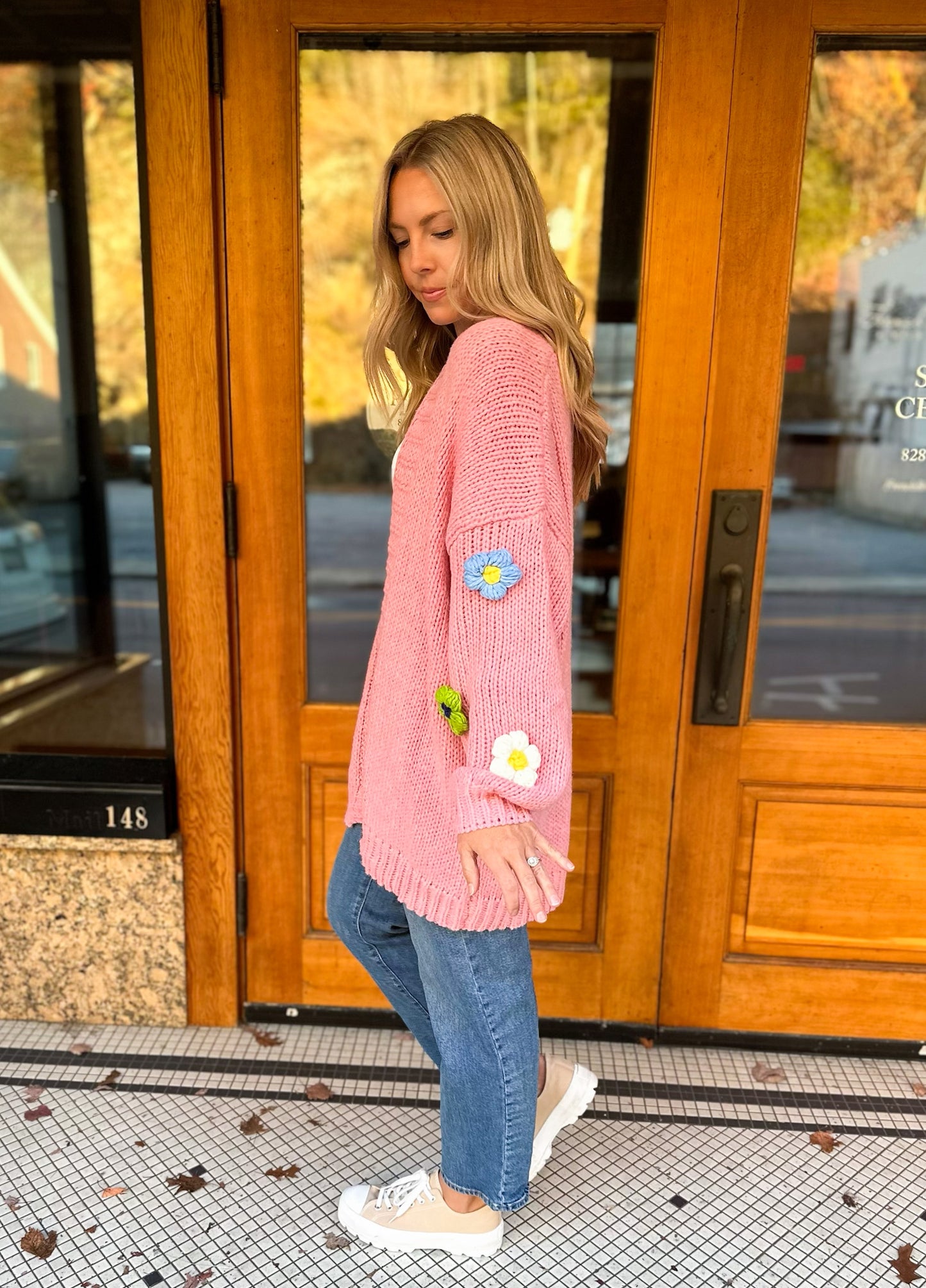 Pink crochet cardigan with an oversized fit with 3-D flowers of various colors on the sleeves. This photo features the side view of the outfit in front of wooden doors.